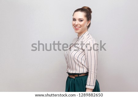 Profile side view portrait of happy beautiful young woman in striped shirt, green skirt with makeup and collected ban hairstyle, looking at camera and smiling. Studio shot, isolated on grey background