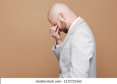 Profile side view portrait of sad crying middle aged bald bearded businessman in light gray suit standing and closed eyes with hands. indoor studio shot, isolated on light brown background.