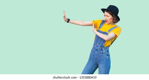 Profile Side View Portrait Of Scared Young Hipster Girl In Blue Denim Overalls, Yellow Shirt And Black Hat Standing With Stop Hand Sign Gesture. Indoor Studio Shot Isolated On Light Green Background.