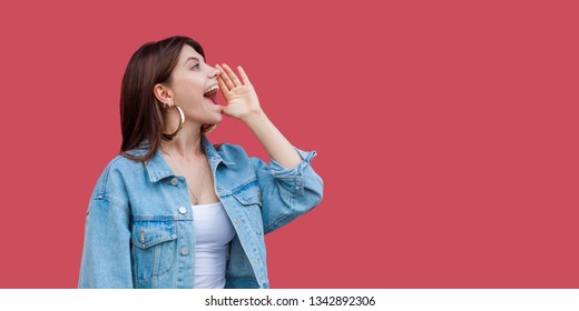 Profile Side View Portrait Of Screaming Beautiful Brunette Young Woman With Makeup In Denim Casual Style Standing With Hand, Shouting At Copyspace Wall. Indoor Studio Shot, Isolated On Red Background.