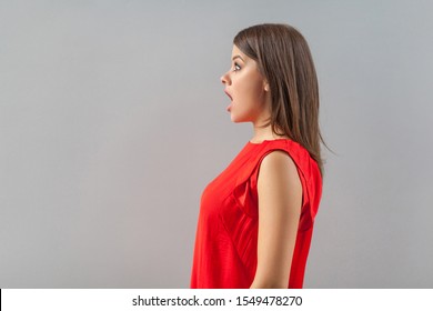 Profile Side View Portrait Of Shocked Beautiful Brunette Young Woman In Red Shirt Standing, Looking Forward With Surprised Unbelievable Face. Indoor, Studio Shot, Isolated On Gray Background.