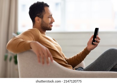 Profile side view portrait of young smiling Arab man holding mobile phone sitting on the couch in living room. Cool guy browsing internet, surfing web or watching video, using application, free space