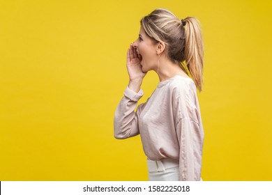 Profile Side View Of Positive Beautiful Blonde Woman With Ponytale Hairstyle And In Casual Beige Blouse Standing With Hand Over Mouth And Shouting. Indoor Studio Shot Isolated On Yellow Background