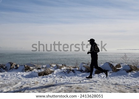 a profile silhouette of a person dressed in black jogging on a frigidly cold winter day on the snow covered rocky shore of a great lake 