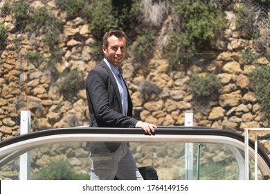 Profile Of A Smiling Businessman Standing At The Top Of An Escalator In The Middle Of The Street And In Front Of A Stone Wall