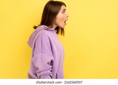 Profile Of Surprised Dark Haired Woman Looking With Shocked Expression To Side, Keeps Mouth Widely Open, Wearing Purple Hoodie. Indoor Studio Shot Isolated On Yellow Background.