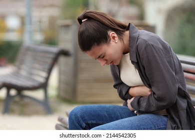 Profile Of A Teenage Female Suffering Belly Ache Sitting On A Bench In A Park