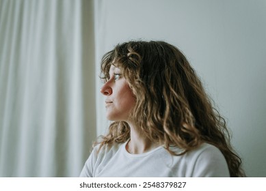 Profile of a thoughtful young woman with curly hair, gazing towards a window with a white curtain, lost in contemplation or daydreaming - Powered by Shutterstock