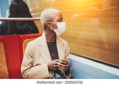 Profile View Of An elegant African Lady In A Facial Anti-virus Protective Mask Sitting In A Car Of A Metro Train With A Smartphone In Her Hands And Looking Outside The Window At The Motion