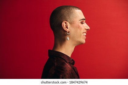 Profile view of androgynous man wearing an earring and makeup smiling. Gay man smiling against red background.