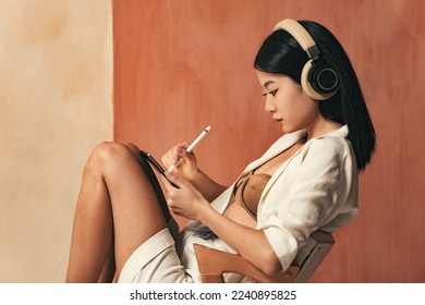 Profile view of asian woman in headphones drawing on tablet with stylus pen, sitting on chair, graphic designer working as freelancer, creating digital art - Shutterstock ID 2240895825