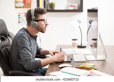 Profile View Of An Attractive Young Man Doing Some Design Work With A Pen Tablet And A Desktop Computer While Listening To Music With Headphones