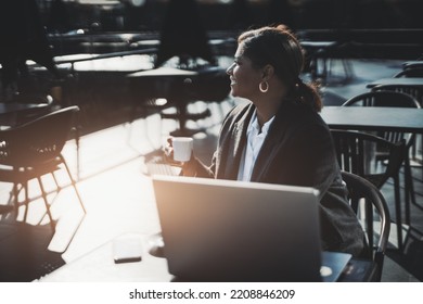 A Profile View Of A Backlit Silhouette Of A Mature Beautiful Hispanic Woman Entrepreneur Sitting In A Street Cafe With Her Laptop While Having Her Coffee Break; A Copy Space Place On The Left