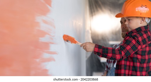 Profile View Of A Boy In A Hard Hat With Dad Painting The Wall In The Room In Orange. The Family Is Doing Home Repairs. Men's Work And Colored Wall. Place For Text. High Quality Photo