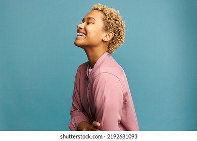 Profile view of cheerful blond african american millennial woman with perfect sense of humor laughing out loud on blue studio background after hearing funny joke. Positive emotions
