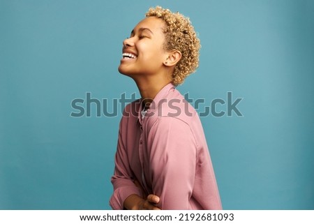 Profile view of cheerful blond african american millennial woman with perfect sense of humor laughing out loud on blue studio background after hearing funny joke. Positive emotions