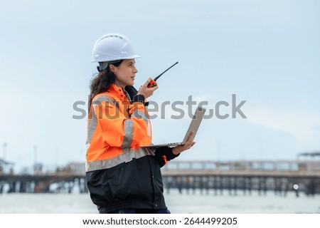Profile view of a female engineer reporting status via radio at a logistics and shipping terminal. Woman supervisor communicating with her team, holding a laptop at a coastal industrial site.