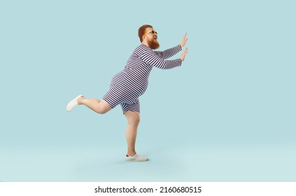 Profile view of funny fat ginger man wearing striped swimsuit and sunglasses dancing and having fun isolated on light blue colour background. Summer holiday, vacation, beach party concept