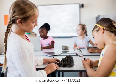 Profile View Of Girls Looking At Rock With Magnifying Glass In Classroom