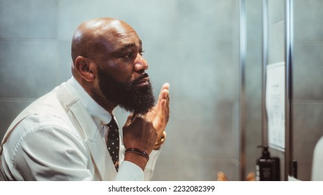A profile view of a handsome bald black man grooming, brushing, and moisturizing the beard hair in front of the mirror in a bathroom of a luxury hotel; an African-American man taking care of his beard