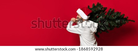 Profile view of happy modern Santa Claus in red hat, glasses and white hoodie carries a Christmas tree on his shoulders on a red background. Winter holiday celebration concept. Place for text. banner