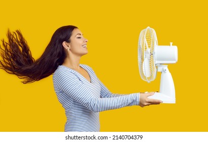 Profile View Of Happy Young Woman Standing Isolated On Yellow Background, Holding Electric Fan, Enjoying Cool Wind Blowing In Her Face, Breathing In Fresh Air And Smiling With Her Eyes Closed