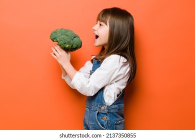 Profile View Of A Little Brunette Girl Eating Some Broccoli In A Studio With Her Mouth Wide Open