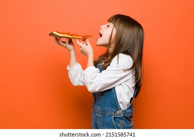 Profile view of a little brunette girl dressed casually and about to bite a slice of pizza in a studio