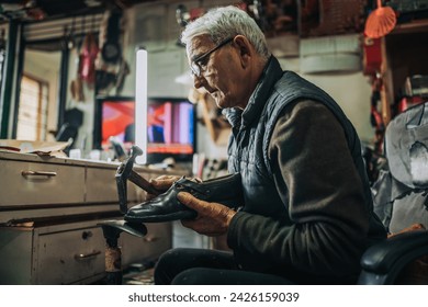 Profile view of an old handyman repairing shoes at shoemaker workshop. A senior cobbler is sitting at his small workshop and fixing shoes and using hammer. A craftsman is repairing shoes at workshop. - Powered by Shutterstock