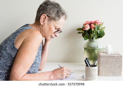 Profile View Of Older Woman With Short Grey Hair And Glasses Sitting At Desk Writing (selective Focus)