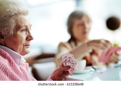 Profile View Of Pensive Elderly Woman With Deep Blue Eyes Sitting At Table And Playing Cards With Her Female Friend