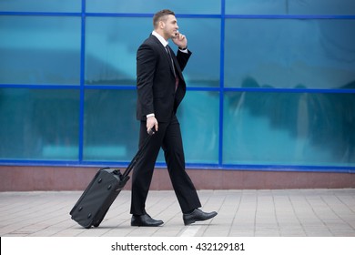 Profile View Portrait Of Cheerful Young Man On Business Trip Walking With His Luggage While Talking On Smartphone In Front Of Modern Glass Building Outdoors. Travelling Guy Making Call. Copy Space