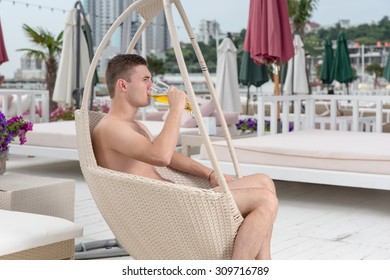 Profile View Of Relaxed Young Man Sitting In Hanging Wicker Chair Drinking Refreshing Glass Of Beer On Luxury Ocean Front Resort Patio Deck