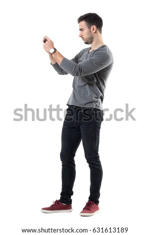 Profile view of serious young casual man holding cellphone taking photo. Full body length portrait isolated over white studio background.
