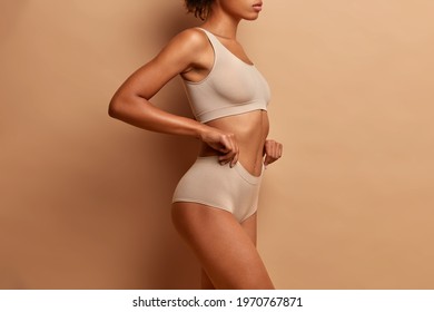 Profile view of slim dark skinned woman dressed in cropped top and panties demonstrates her perfect body slender legs poses against brown background