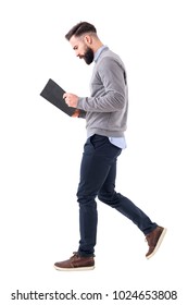 Profile view of young bearded businessman walking while reading notebook or planner. Full body length portrait isolated on white studio background.