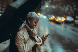 A Profile View Of A Young Elegant Cheerful Black Lady With Short Painted White Hair, Using Her Umbrella On The Rainy Street While Trying To Call A Taxi Via An App On Her Smartphone To Escape The Rain