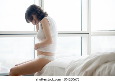 Profile View Of Young Pregnant Woman Sitting On The Bed Covering Her Mouth Feeling Nauseous In Early Stages Of Pregnancy. Morning Sickness