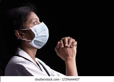 Profile View Of Young Woman Doctor In Medical Mask Praying To God On Black Background Looking Up - Concept Of Hope And Fight To End Coronavirus Or Covid-19 Crisis.