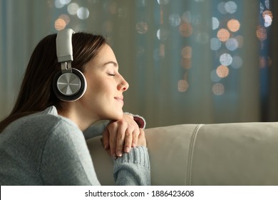 Profile Of A Woman With Closed Eyes Feeling And Listening To Music With Wireless Headphones Sitting On A Couch In The Night At Home