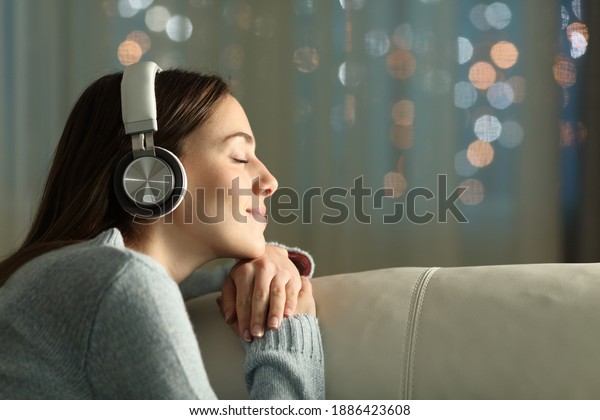 Profile of a woman with closed eyes feeling and\
listening to music with wireless headphones sitting on a couch in\
the night at home