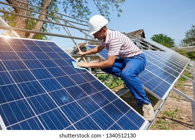 Profile of worker in white helmet wiping clean big square shiny solar panel standing on tall metal platform on summer bright blue sky background. Renewable energy generation concept. - Shutterstock ID 1340512130