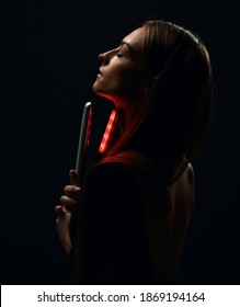 Profile of young beautiful woman with long silky straight hair in black body with eyes closed holding hair straightener in hand over dark background. Haircare, beauty, wellness concept