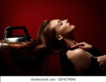 Profile of Young beautiful woman posing with eyes closed and her long silky straight hair under iron on board over dark red background. Haircare, beauty, wellness, hairstyle, straightening concept