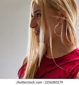 Profile of young blond woman with mouth half-open, in red dress, with a heart-shaped earring in her ear, looking intently at something or someone and smiling slightly. - Shutterstock ID 2256383179