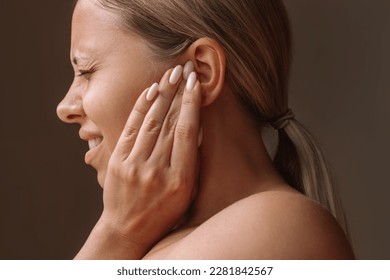 Profile of a young caucasian blonde woman holding her ears with her hands isolated on a dark brown background. Pain and tinnitus, ear otitis