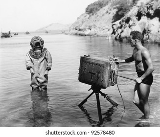 Profile of a young man holding a camera with a scuba diver standing in front of him on the beach - Powered by Shutterstock