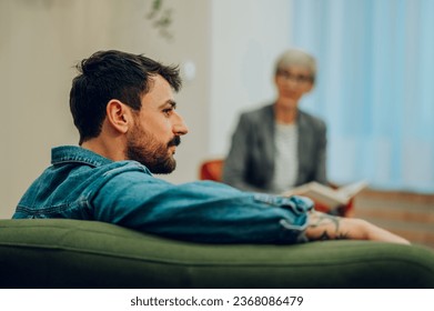 Profile of a young man with issues sitting on a sofa in the office with a psychotherapist and discussing trauma and problems. In a blurry background is the therapist giving support and giving advice.