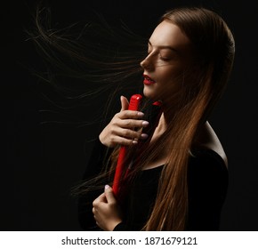 Profile of young woman with long silky fluttering in the wind hair standing with eyes closed holding red hair straightener at face over dark background. Haircare, beauty, wellness concept