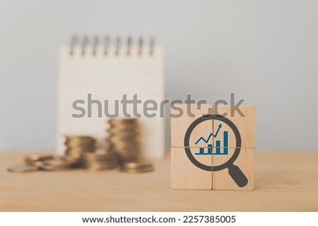 For profitability concept. Business profit results; sales, margin, cost management.  wooden cubes with blue increasing graph inside grey magnifying glass icon and blurred stack of coins and calendar